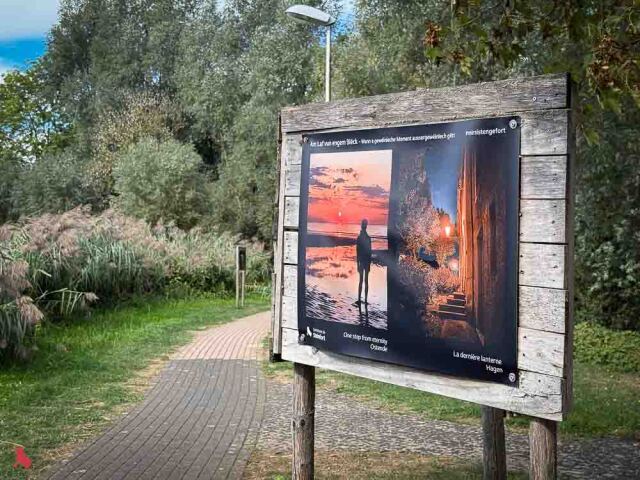 Panneau de l'exposition Billerwee « Am Laf vun engem Bléck » au bord d'un chemin, avec les photos « One step from eternity » (Ostende) et « La dernière lanterne » (Hagen)
