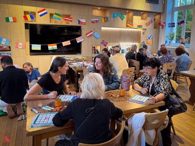 Des participant·e·s discutent par petits groupes autour de grilles de jeu, sous des guirlandes de drapeaux, à l'Afterwork interculturel
