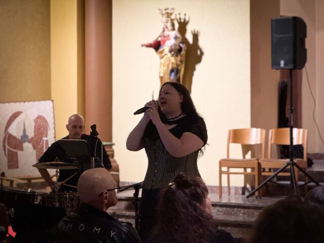 La chanteuse du groupe ShadoWhisperS chante au micro devant le public, avec le batteur derrière elle, lors du concert « Metal & Pipes »