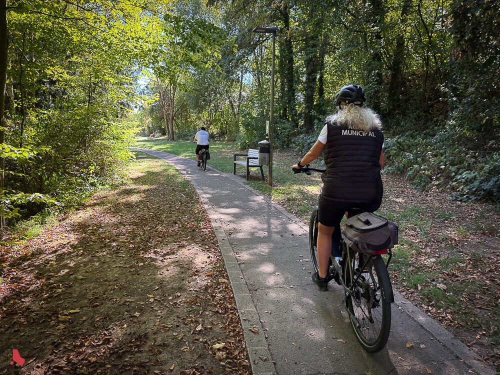 Deux agent·e·s municipaux·ales à vélo sur un chemin boisé couvert de feuilles mortes