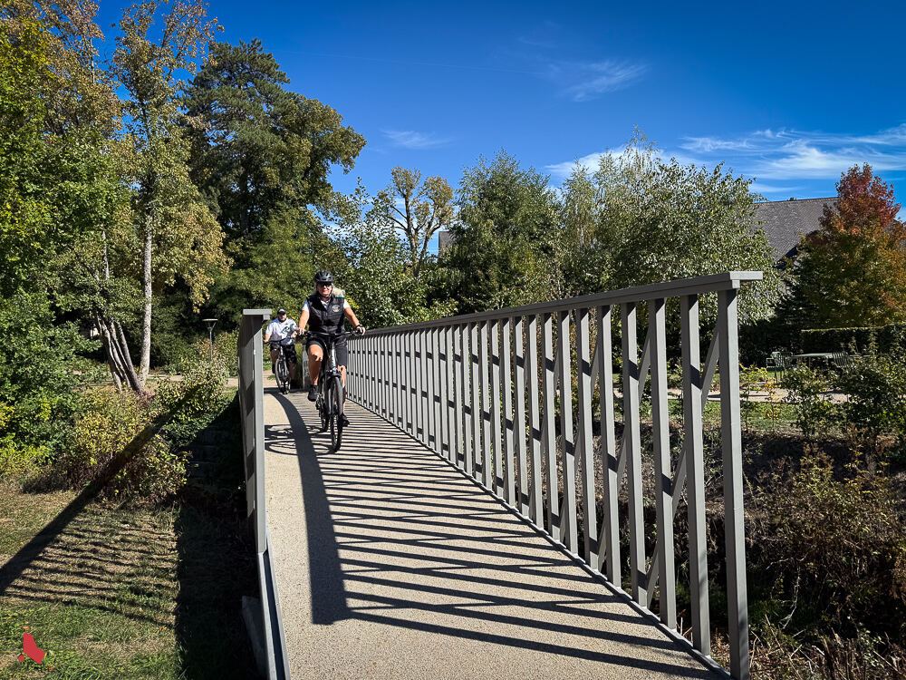 Deux agent·e·s municipaux·ales à vélo sur une passerelle dans un parc arboré