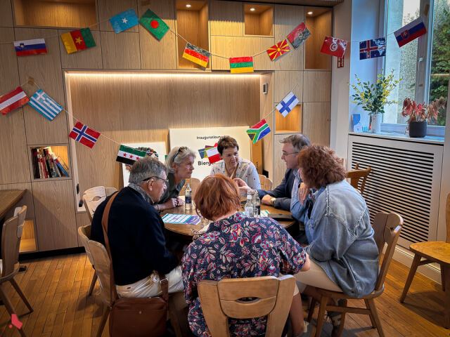 Six participant·e·s discutent autour d'une table sous des guirlandes de drapeaux du monde, à l'Afterwork interculturel au Bierger*Haus