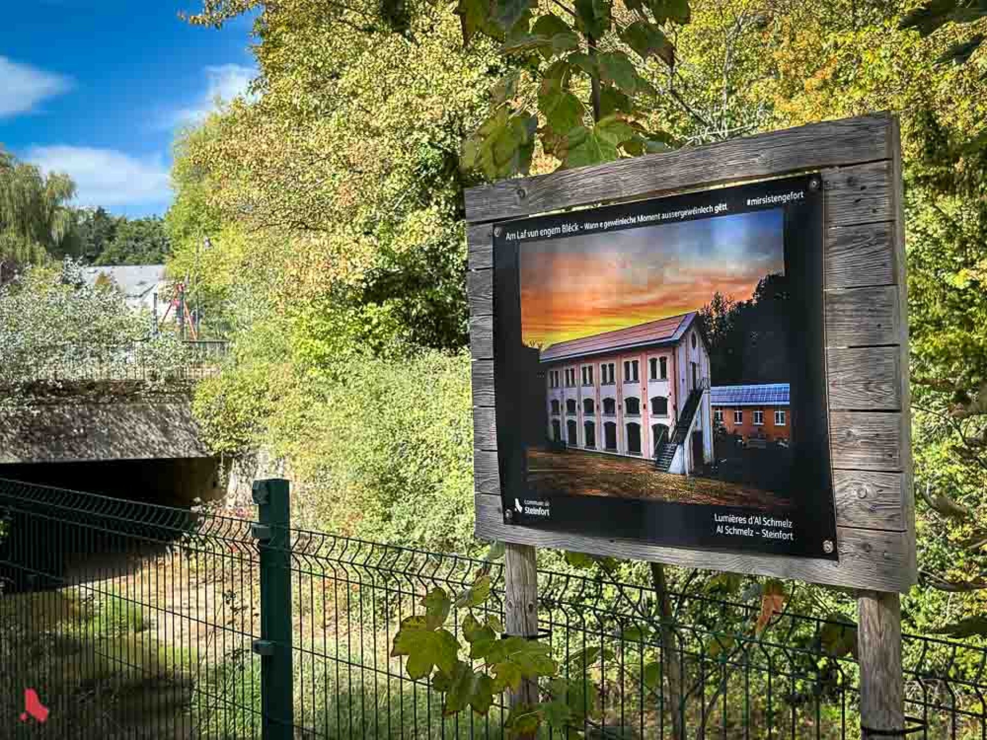 Panneau de l'exposition Billerwee « Am Laf vun engem Bléck » avec la photo « Lumières d'Al Schmelz », le bâtiment d'Al Schmelz au coucher du soleil