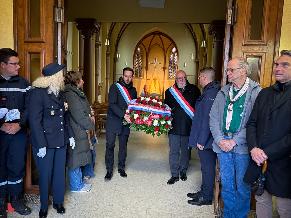 Deux élus en écharpe tricolore portent une gerbe à l’entrée de l’église, lors de la Journée de commémoration nationale