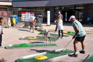 Adultes et enfants jouent au minigolf sur des pistes mobiles installées sur la place devant le centre sportif